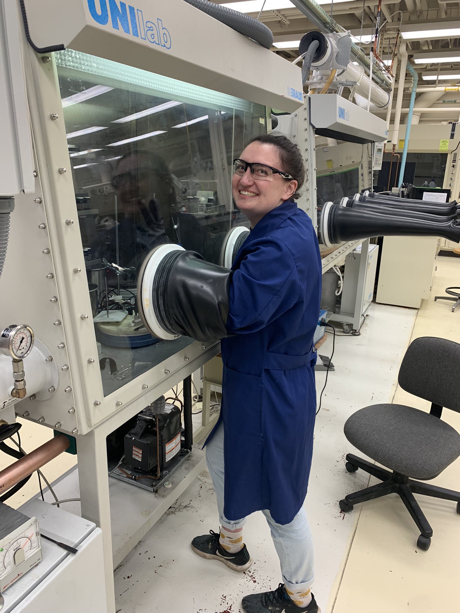 Becca Segel smiling in a laboratory wearing a dark blue lab coat and safety glasses.